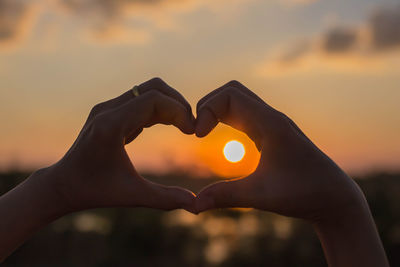 Woman making heart shape in front of sun against sky during sunset