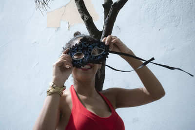 Portrait of a woman wearing a carnival mask against a light blue wall.