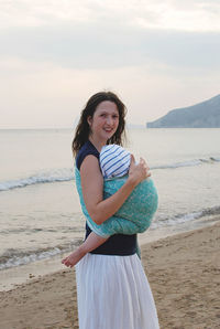 Portrait of young woman holding baby wrapped in baby blanket standing at beach