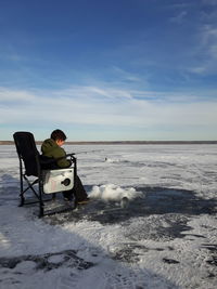 Boy ice fishing while sitting on chair against blue sky