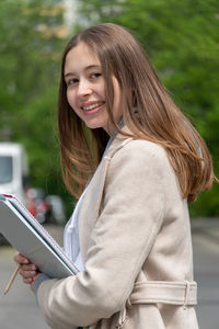 Portrait of smiling teenage girl holding books against trees