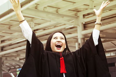 Cheerful young woman in graduation gown with arms raised 