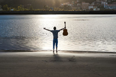 Back view of unrecognizable male musicians standing with acoustic guitar in outstretched arms on wet beach and enjoying freedom