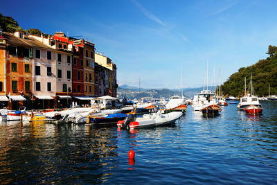 Boats moored in sea by buildings against blue sky