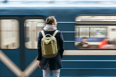 Handsome man in a blue coat with green backpack behind standing on subway platform, train in motion.