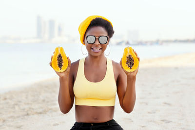 Cheerful african girl in sunglasses sitting on the sand, holding a papaya, smiling and posing.