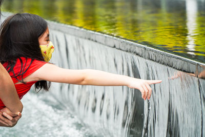 Cute girl wearing mask touching flowing water outdoors