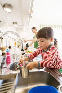 Little girl filling water in container with mother in background at kitchen counter