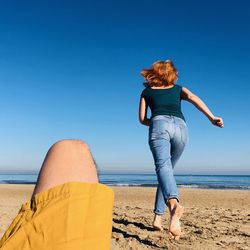 Rear view of mid adult woman running on beach against clear blue sky during sunny day