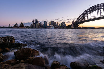 Bridge over sea by buildings against sky during sunset