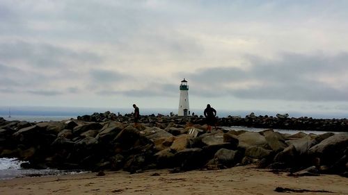 Men on groyne at beach against cloudy sky