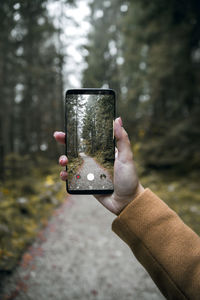 Cropped hand of woman photographing road amidst trees in forest