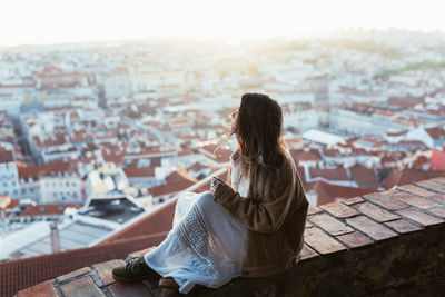 Side view of young lady in jacket and dress sitting on brick barrier and admiring cityscape in sunny morning in portugal