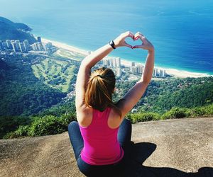 Rear view of woman with arms raised making heart shape on mountain cliff