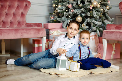 Portrait of cute sibling sitting by christmas tree at home
