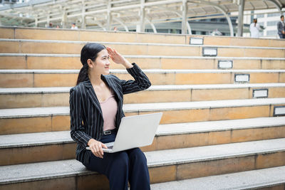 Smiling young businesswoman using laptop on steps in city