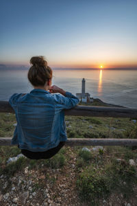 Rear view of woman sitting on railing while looking at sea against sky during sunset