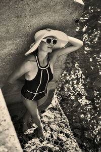 Portrait of a woman in a swimsuit, hat and sunglasses in summer on the riverbank by a concrete wall