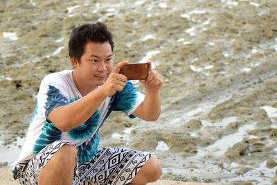 High angle view of young man photographing with mobile phone while crouching at beach