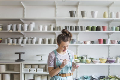 Young female potter using hand tool on vase while standing against shelves at store