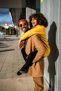 Cool african american man in sunglasses and coat giving piggyback ride to bright laughing girlfriend with afro curly hair spending time on street