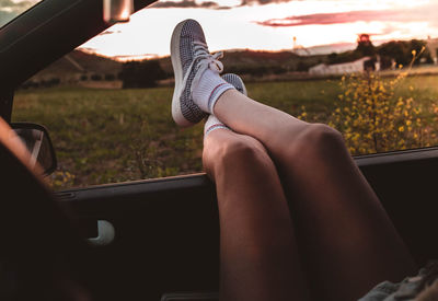 Low section of woman relaxing on car window against sky during sunset