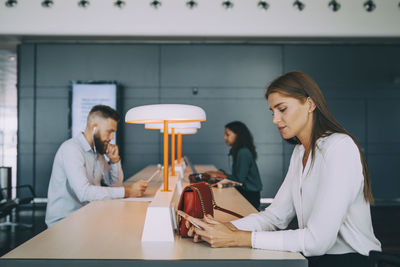 Young businesswoman using smart phone while sitting with multi-ethnic colleagues at table waiting in airport departure a