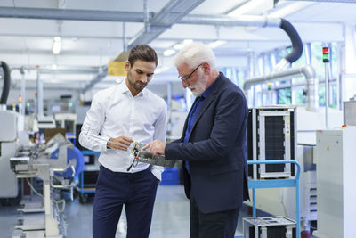 Senior manager pointing at machine part while discussing with engineer by machinery in factory