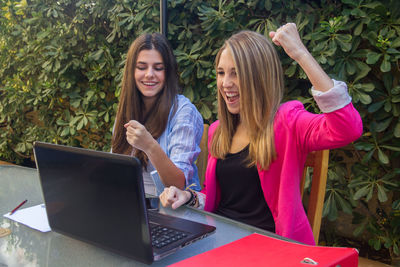 Female colleagues clenching fists while using laptop against plants