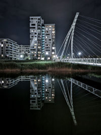 Illuminated bridge over river by buildings against sky at night