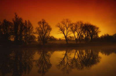 Silhouette trees by lake against orange sky
