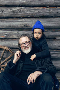 Grandson in a blue hat and boots with a grandfather in a sheepskin coat stand at a wooden house shed