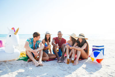 Friends holding drinks while sitting at beach against sky