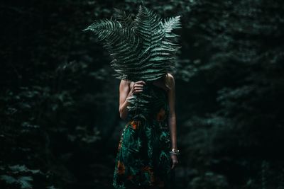 Woman covering face with leaves while standing in forest