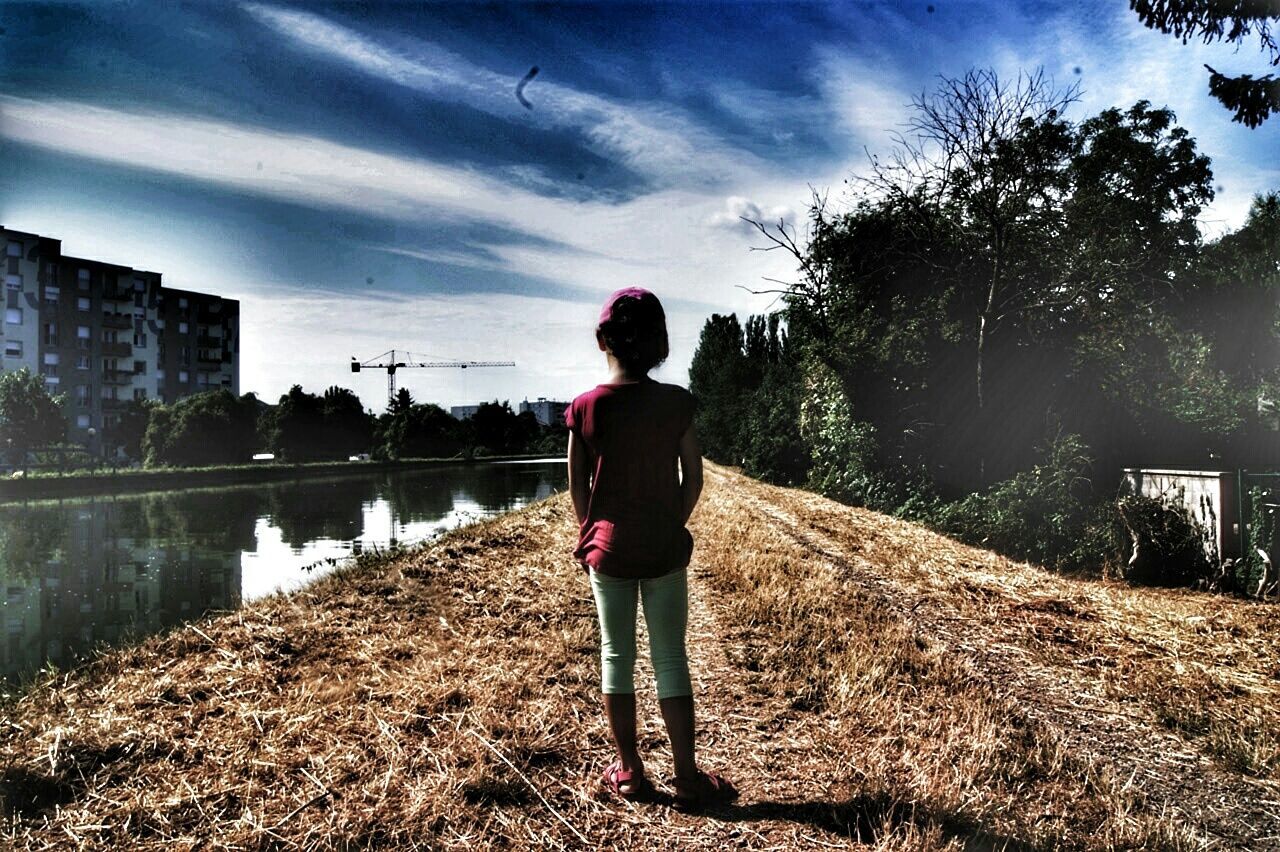 water, full length, sky, lifestyles, standing, rear view, building exterior, built structure, casual clothing, architecture, reflection, cloud - sky, leisure activity, walking, cloud, tree, river, day