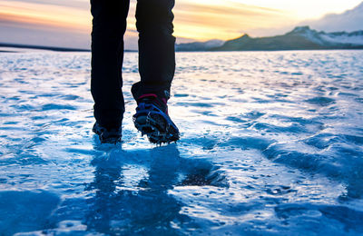 Low section of man walking frozen lake against sky during sunset