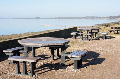 Empty benches and table on shore against sky