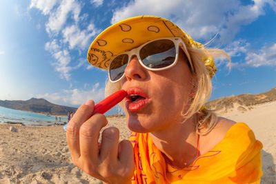Woman applying lipstick at beach against sky