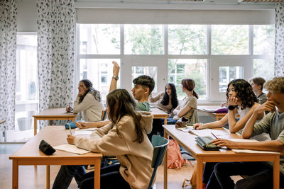 Boy raising hand while sitting with other students in classroom