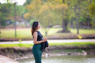 Side view of smiling young woman standing by lake and trees