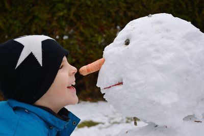 Side view of cheerful boy standing by snowman