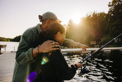 Male counselor gesturing by girl fishing on jetty near lake at summer camp