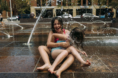 Cheerful sisters wearing swimwear playing with fountain water against buildings in city