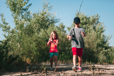 Full length of siblings with fishing rods walking on field