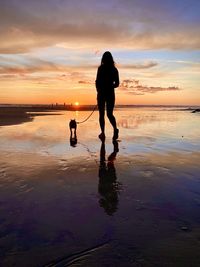 Silhouette woman with dog walking at beach against sky during sunset