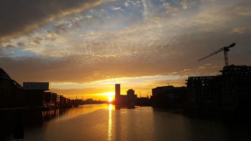 Silhouette buildings by river against sky during sunset