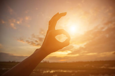 Cropped hand of woman gesturing ok sign against cloudy sky during sunset