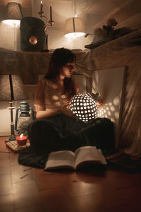 Serene female in domestic wear sitting in cozy room in evening with illuminated vintage lantern while chilling at home and looking away
