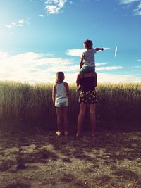 Rear view of woman piggybacking son pointing while standing by daughter on land against sky