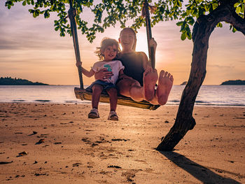 Full length of woman and child on beach swing against sky during sunset